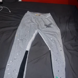 Grå mjukisbyxor från Gallery Dept. - Grå mjukisbyxor från Gallery Dept. med svart och vit detalj längs benen och tryckt logga på ena låret. Byxorna har resår i midjan och ett avslappnat snitt. Färgade stänk och slitna detaljer ger en unik look.
