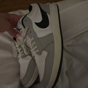 Nike Air Jordans i vitt och grått - Säljer ett par Nike Air Jordans med vit bas, ljusgråa paneler och svart swoosh. Skorna har snörning, perforerad tå och klassisk Air Jordan-logga på hälen. Perfekta för dig som gillar streetwear och sneakers med ikonisk design.