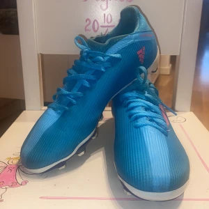 Blå adidas fotbollsskor med rosa detaljer - Säljer ett par blå adidas fotbollsskor med rosa logga och detaljer. Skorna har snörning och är tillverkade i syntetmaterial med mönstrad ovandel. Perfekta för dig som vill sticka ut på planen!