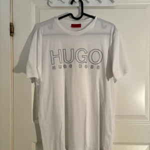 Vit t-shirt från Hugo Boss - Säljer en vit t-shirt från Hugo Boss med stor logga i silvergrått framtill. Klassisk rund halsringning och korta ärmar. Perfekt för en stilren och enkel look.