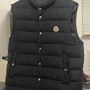 Svart dunväst från Moncler - Säljer en svart dunväst från Moncler med klassisk quiltad design och tryckknappar framtill. Västen har två fickor med knappstängning och Moncler-logga på bröstet. Perfekt för sommaren storlek 2 