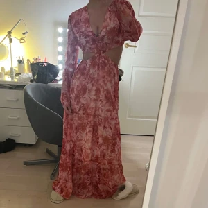 klänning - Säljer en maxiklänning i rosa och röda toner med blommigt mönster. Klänningen har lång ärm, djup v-ringning och trendig cut-out i midjan. Flödande material och snygg passform, perfekt för sommarens tillfällen.