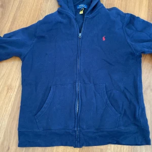 Mörkblå hoodie med dragkedja från Polo Ralph Lauren - Mörkblå hoodie från Polo Ralph Lauren med dragkedja framtill och två fickor. Klassisk röd logga broderad på bröstet. Perfekt för en avslappnad stil och enkel att matcha med andra plagg. Tröjan passar de som är 163-174cm