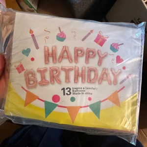 Happy Birthday ballongkit - Säljer ett ballongkit med texten 'Happy Birthday' i rosa ballonger, samt färgglada vimplar och dekorationer med tårtbitar och presenter. Perfekt för att dekorera till födelsedagsfesten. Innehåller ballonger och tillbehör.