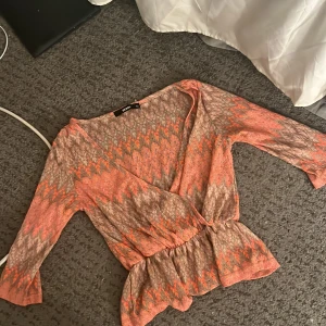 Mönstrad omlottopp från Bik Bok - Säljer en snygg omlottopp från Bik Bok i storlek XS. Toppen har ett orange och beige zigzag-mönster, v-ringning och trekvartsärm. Resår i midjan ger en fin passform och toppen är i ett lätt, syntetmaterial.