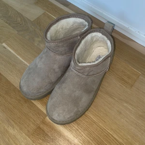 Beige boots från Boohoo med foder - Mysiga beige boots från Boohoo med mjukt vitt foder. Skorna har rund tå, platt sula och är tillverkade i syntetmaterial med mockaliknande yta. Perfekta för kyligare dagar och enkel att dra på tack vare den låga modellen.