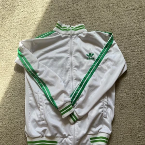 Vit Adidas track jacket med gröna ränder - Klassisk vit Adidas track jacket med gröna ränder längs ärmarna och gröna detaljer vid krage, ärmslut och midja. Jackan har dragkedja och Adidas logga både fram och bak. Perfekt för en sportig och retro stil.