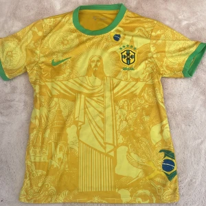 Brasilien fotbollströja  - Gul Brasilien fotbollströja med gröna detaljer från Nike. Framsidan har ett stort motiv av Kristusstatyn i Rio och brasilianska symboler. Broderad landslagslogga och Nike-logga på bröstet. Rund hals och korta ärmar.
