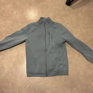 Ljusblå jacka från Sail Racing - Säljer en ljusblå jacka från Sail Racing med hög krage och hel dragkedja framtill. Jackan har en bröstficka med dragkedja och Sail Racing-logga broderad på bröstet. Mjuk insida och stilren design.