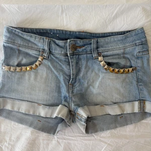 Lågmidjade jeansshorts med nitar - Säljer ett par skitsnygga lågmidjade ljusblå jeansshorts med snygga nitar vid fickorna. 38 cm i midjemått. Shortsen har uppvikta benslut och klassisk femficksdesign. Perfekta för sommaren!☀️✨