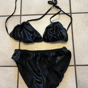 Svart glittrig bikini med knyt - Snygg svart bikini med glittrigt material och trekantsöverdel med knyt i nacken och ryggen. Underdelen har hög midja och samma glittriga finish. Perfekt för dig som vill sticka ut på stranden. Storlek XS/S