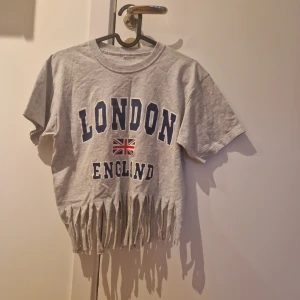 Grå London t-shirt med fransar - Grå kort t-shirt med tryck 'LONDON ENGLAND' och brittisk flagga på bröstet. T-shirten har korta ärmar och är croppad med fransar längst ner för en cool och avslappnad stil. Perfekt för dig som gillar streetwear och unika detaljer.