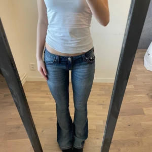 Blå bootcut jeans från True Religion - Säljer ett par blå jeans från True Religion med bootcut passform och låg midja. Jeansen har lila röda sömmar och loggan på fickorna. Jag är 164 för referens! Innerbenslängden är 82 cm och Midjemått är 36 cm. 