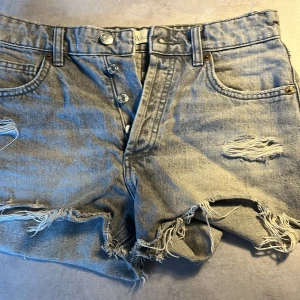 Ljusblå jeansshorts med slitningar och knappar - Ljusblå jeansshorts med slitna detaljer och fransiga kanter. Klassisk femficksmodell med knappar framtill och bälteshällor. Perfekta för en avslappnad sommarstil.