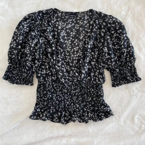 Svart blommig blus från KappAhl - Supersöt svart blus med vita småblommor från KappAhl. Blusen har v-ringning, volangdetaljer och smockad midja samt ärmar med puff och resår. Perfekt för vår och sommar!