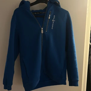 Blå hoodiejacka från Sail Racing - Blå hoodiejacka från Sail Racing med dragkedja framtill och på bröstficka. Jackan har huva och vit logotyp på bröstet. Perfekt för en sportig stil och enkel att matcha med jeans eller träningsbyxor.