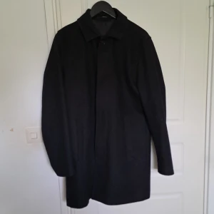 Svart rock från Tiger of Sweden - Stilren svart rock från Tiger of Sweden med klassisk krage och raka fickor. Perfekt för dig som vill ha en tidlös och elegant look. Fodrad insida för extra komfort.