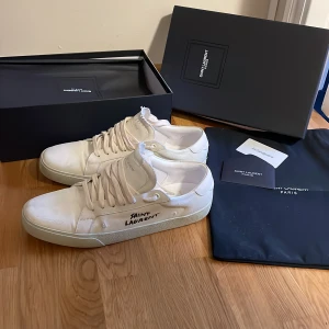 Vita Saint Laurent sneakers - Säljer ett par vita sneakers från Saint Laurent med svart logga på sidan. Använda ett fåtal gånger, som nyskick. Klassisk och stilren design i canvas och skinn. Kommer med originalkartong och dustbag. 