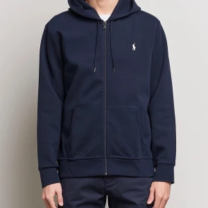 Mörkblå hoodie med dragkedja från Polo Ralph Lauren - (OÖPPNAD MED KVITTO) Säljer en mörkblå hoodie från Polo Ralph Lauren i storlek L. Tröjan har dragkedja, huva med snören och två fickor framtill. Klassisk broderad logga på bröstet. Perfekt för en avslappnad stil.