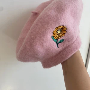 Rosa basker med broderad blomma från Minimarket - Söt rosa basker från Minimarket i ull med en broderad gul blomma på sidan. Klassisk form med liten toppknopp. Perfekt accessoar för att lyfta din outfit med en färgklick och unik detalj.