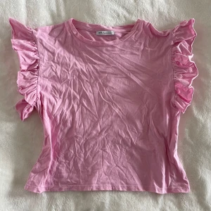 Rosa volangtopp från Zara - Säljer en söt rosa topp från Zara med volangdetaljer på axlarna. Toppen har rund halsringning och är tillverkad i mjuk bomull. Perfekt för dig som gillar feminina detaljer och en poppig färg i garderoben!
