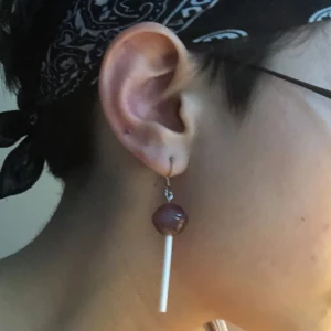Örhänge med klubba - Unikt örhänge format som en rödbrun klubba med vit pinne. Perfekt för dig som vill sticka ut och ha något lekfullt i örat. Fäste i metall och cool design som drar blickarna till sig.