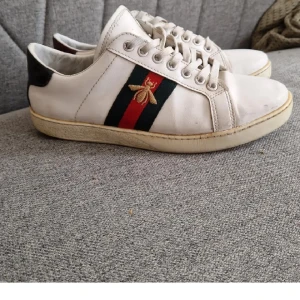 Gucci Ace sneakers med bi-detalj - Säljer ett par vita Gucci Ace sneakers med klassiska gröna och röda ränder samt broderad guldfärgad bi på sidan. Baksidan har ormskinnsmönster i rött och svart. Skorna har snörning och platt sula. Nypris ca 8500. Tar byten!