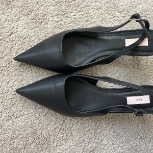 Svarta pumps med spetsig tå från Nelly - Säljer ett par stilrena svarta pumps från Nelly med spetsig tå och smal klack. Skorna har en öppen häl och smala remmar som går runt ankeln. Tyvärr aldrig använda då dom inte passade mig i storleken och har nu gått för lång tid för att lämna tillbaka.
