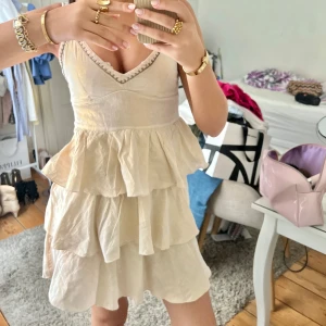 Champagnefärgad/rosa volangklänning med v-ringning - Säljer en söt champagnefärgad/rosa klänning med volanger och v-ringad hals. Klänningen är ärmlös och har en omlottliknande överdel med fina spetsdetaljer längs urringningen. Perfekt för sommarens alla tillfällen!