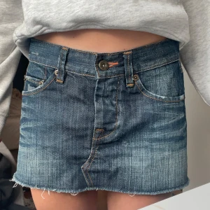 Jeanskjol i mini-modell - Säljer en klassisk blå jeanskjol i mini-längd med råa kanter och femficksdesign. Kjolen har synliga sömmar i kontrastfärg och knappstängning framtill. Perfekt för dig som gillar en avslappnad och trendig stil.