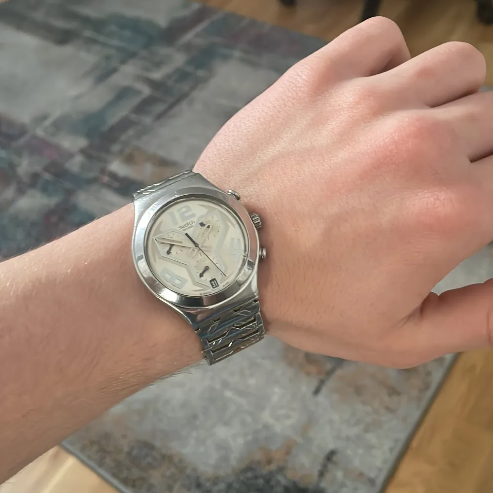 Säljer en stilren silverfärgad klocka från Swatch med ett unikt mönster på armbandet. Klockan har en ljus urtavla med stora siffror och datumvisning. Perfekt för den som gillar en modern och iögonfallande design. Klockans batteri behöver bytas.. Asusteet.