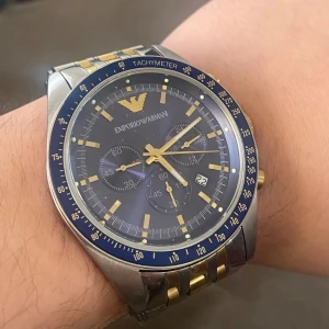 Emporio Armani klocka med blå urtavla - Snygg Emporio Armani klocka med blå urtavla och guldiga detaljer. Klockan har en tachymeter och datumvisning. Armbandet är i tvåfärgat stål och guld, vilket ger en elegant look. Perfekt för den stilmedvetne!