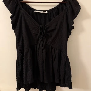 Svart blus med volangdetaljer - Säljer en svart blus med volangdetaljer och knytning framtill. Blusen har korta ärmar och en luftig passform, perfekt för både vardag och fest. Den är tillverkad i ett lätt och bekvämt material.