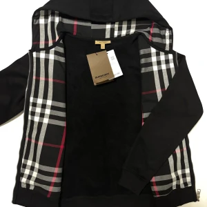 Burberry tröja  - Snygg svart hoodie från Burberry med klassiskt rutigt mönster på insidan av huvan. Den har dragkedja framtill och justerbara snören vid huvan. Perfekt för en stilren look.