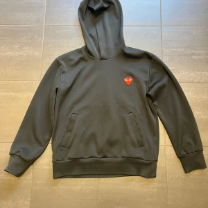 Comme des GARÇONS - Säljer en stilren svart hoodie med ett litet rött hjärta på bröstet. Tröjan har långa ärmar och en klassisk huva. Perfekt för en avslappnad stil.