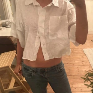 Vit croppad skjorta - Säljer en vit croppad skjorta med korta ärmar och knappar framtill. Skjortan har en bröstficka och är tillverkad i ett lätt och luftigt material, perfekt för varmare dagar.