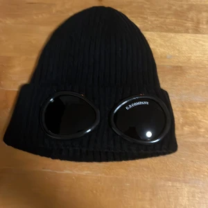 Cp company mössa - Snygg svart ribbad mössa med inbyggda glasögon från C.P. Company. Perfekt för att ge din outfit en unik touch. Mössan är både stilren och funktionell.