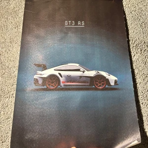 Porsche poster/affisch - Snygg poster med en bild av en vit GT3 RS bil mot en blå bakgrund. Bilen har röda detaljer och en stor bakspoiler. Perfekt för bilentusiaster som vill ha en stilren och sportig väggdekoration.