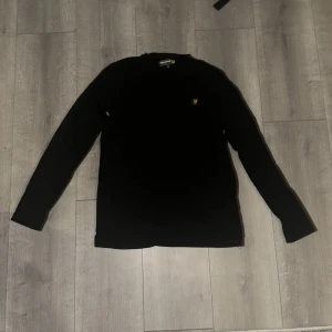 Svart tröja från Lyle & Scott - Säljer en stilren svart tröja från Lyle & Scott med deras ikoniska gula logga på bröstet. Tröjan har långa ärmar och är perfekt för en avslappnad look. Passar bra till både jeans och chinos.