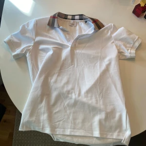 Vit pikétröja från Burberry storlek S/M - Säljer en stilren vit pikétröja från Burberry med den klassiska rutiga kragen i beige, svart och röd. Tröjan har korta ärmar och knappar framtill. Perfekt för en elegant och avslappnad look.