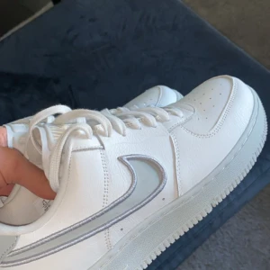 Vita Nike Air Force sneakers - Snygga vita Nike Air Force sneakers med klassisk design och silverdetaljer. Skorna har snörning och en rund tå, vilket ger en bekväm passform. Perfekta för en stilren look.