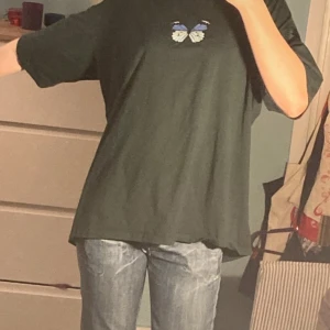 Oversized sommar T-shirt  - Säljer en grön t-shirt med ett snyggt fjärilstryck i blått och vitt på bröstet. T-shirten har en normal passform och korta ärmar, perfekt för en avslappnad stil.