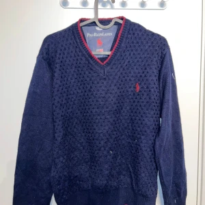 Mörkblå stickad tröja från Polo Ralph Lauren - Säljer en snygg mörkblå stickad tröja från Polo Ralph Lauren med v-ringning och röd broderad logga på bröstet. Tröjan har långa ärmar och en röd kant vid halsen. Perfekt för kyliga dagar!