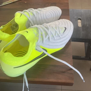 Nike phantom  fotbollsskor i vit och neon - Säljer ett par Nike fotbollsskor i en cool kombination av vit och neon. Skorna har snörning och en rund tå, perfekt för att dominera på planen. De är designade för att ge bra grepp och komfort under spelet.