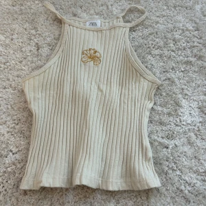 Beige ribbad linnetopp från Zara - Säljer en stilren beige ribbad linnetopp från Zara. Toppen har smala axelband och en broderad blomma framtill. Perfekt för varma dagar eller som ett lager under en jacka.