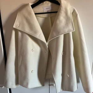 Vit kappa från Bubbleroom - Elegant vit kappa från Bubbleroom med bred krage och knappar framtill. Perfekt för en stilren look under våren. Jackan är använd typ 3 gånger så är väldigt ny, köptes i september. Mycket vitare i verkligheten! 
