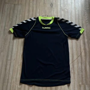 Svart och neongul sporttröja från Hummel - Säljer en sportig svart tröja från Hummel med neongula detaljer och korta ärmar. Tröjan har ett klassiskt Hummel-mönster på ärmarna och är perfekt för träning.