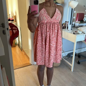 Röd mönstrad klänning - Säljer en röd klänning med vitt paisleymönster. Klänningen är ärmlös med halterneck och har en luftig passform. Perfekt för varma sommardagar!