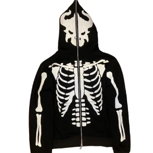 Svart hoodie med skelettmönster - Cool svart hoodie med ett iögonfallande skelettmönster i vitt. Hoodien har en dragkedja framtill och en huva med ett dödskallemotiv. Perfekt för en unik och edgy stil.