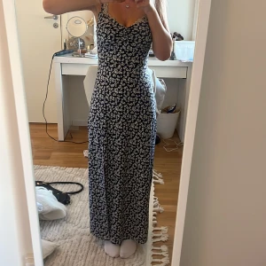 Blommig långklänning med slits - Säljer en super fin långklänning med ett blommigt mönster i marinblå och vitt. Klänningen har smala axelband och en slits framtill. Perfekt till sommaren, säljer den då den är lite för tajt för mig. Tror ej den e använd, möjligtvis en gång
